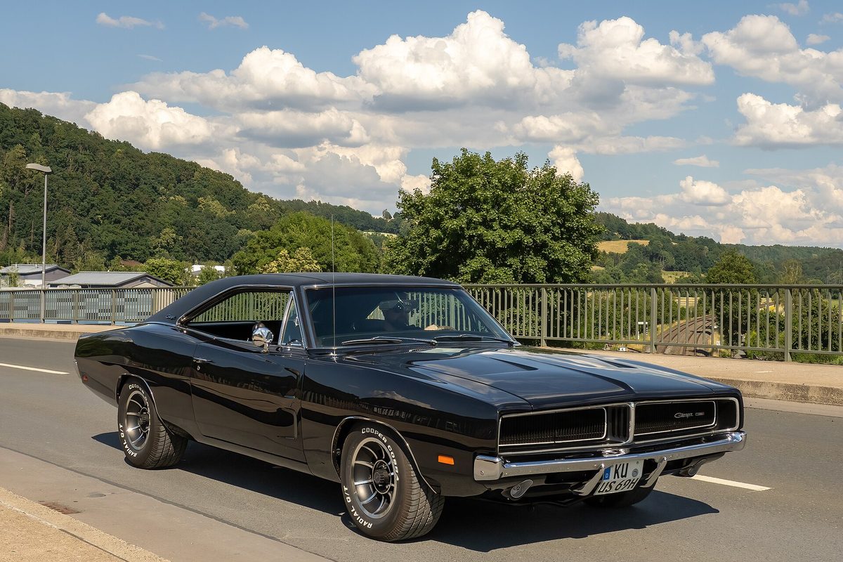 A black 1969 Dodge Charger two-door coupe with chrome bumpers on a bridge road under a cloudy summer sky