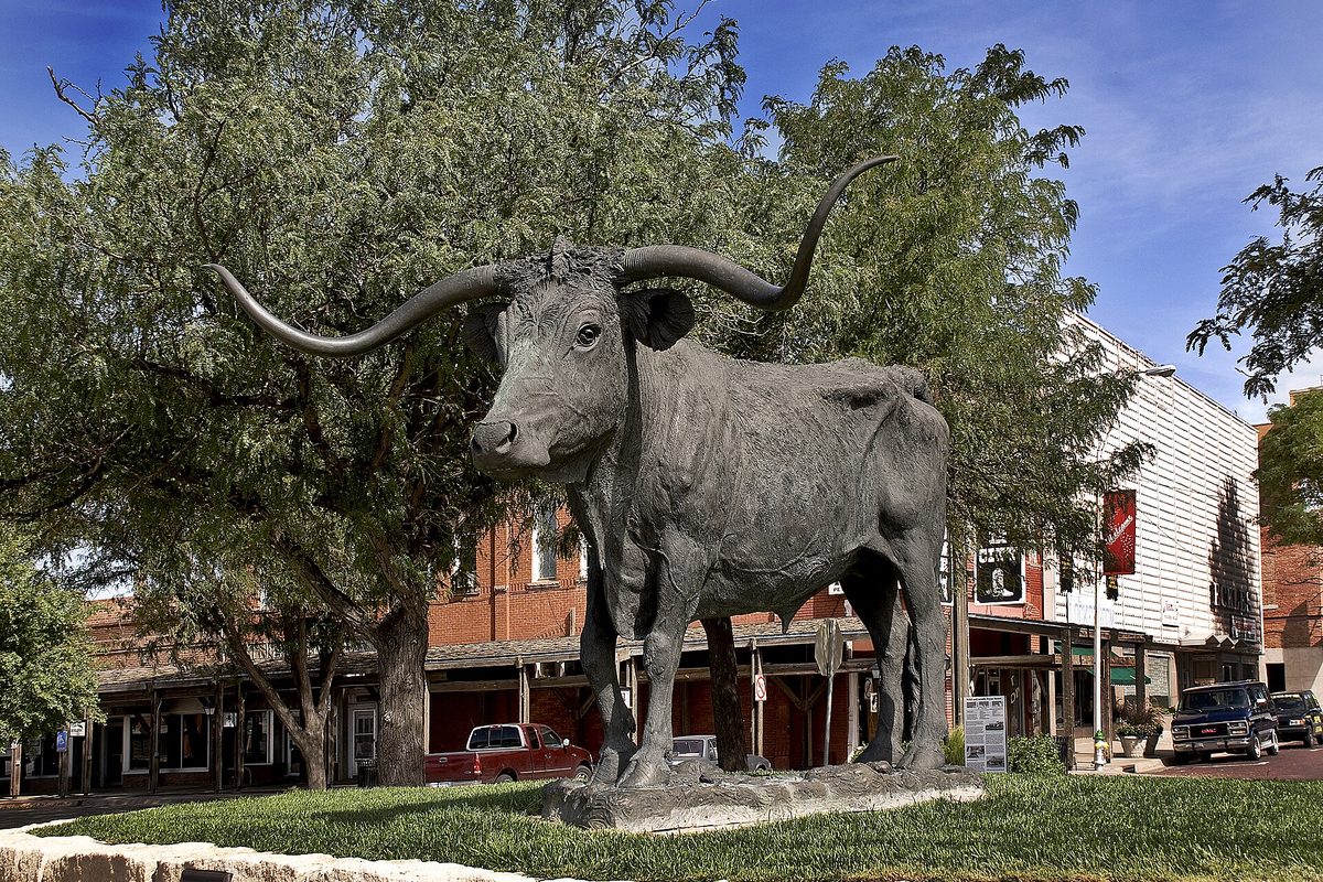 A bronze longhorn steer statue on a grassy island on Front Street in Dodge City, with brick storefronts and parked pickups behind it