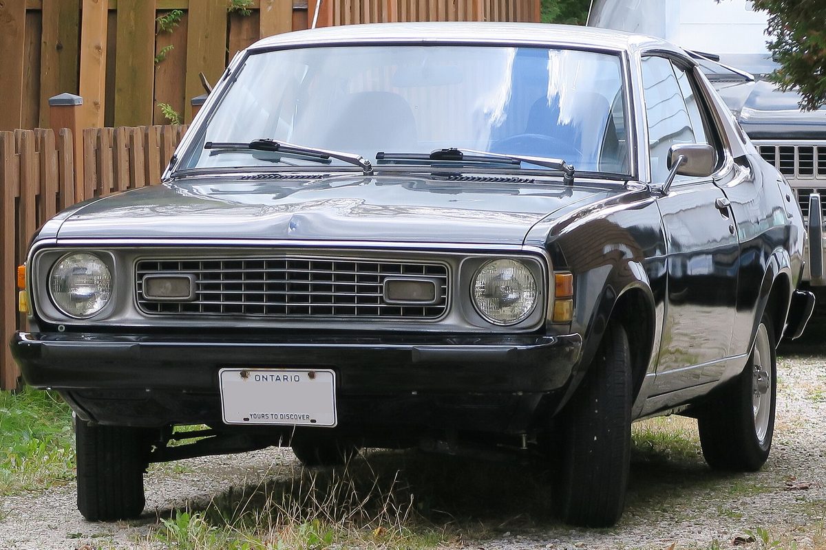 A black second-generation Dodge Colt Carousel two-door hardtop with round headlights, parked on gravel beside a wooden fence