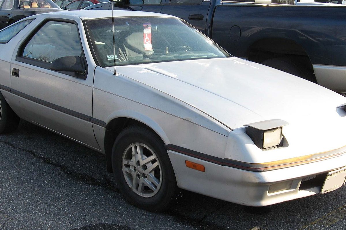 A silver Dodge Daytona hatchback coupe with one pop-up headlight cover raised, parked in a lot next to a pickup