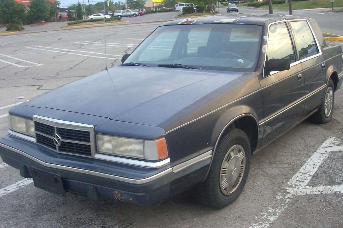 A dark blue 1991-1993 Dodge Dynasty sedan with worn paint, parked alone in an empty lot