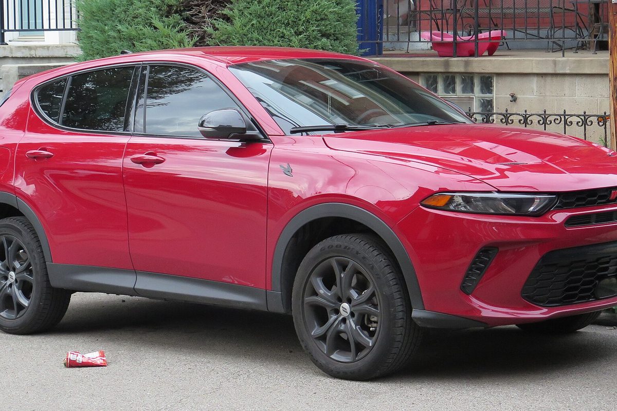 A red Dodge Hornet compact SUV with black wheels, parked on a city street in front of a hedge and an iron fence