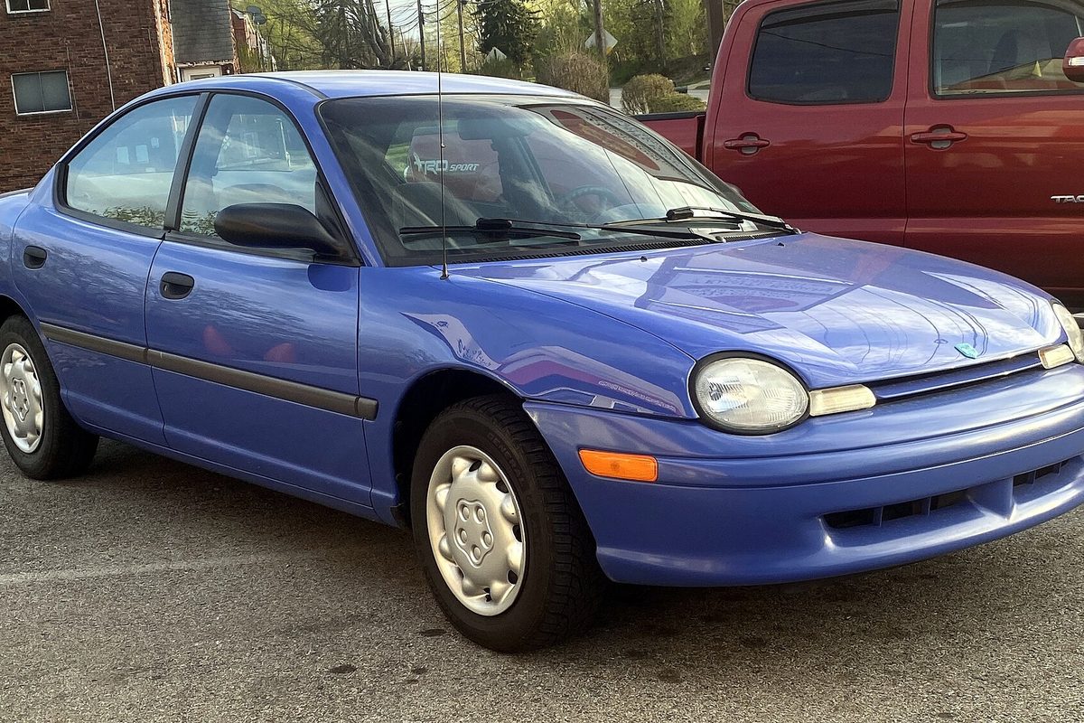 A bright blue 1996 Dodge Neon base sedan with plain hubcaps, parked in a lot in front of a red pickup