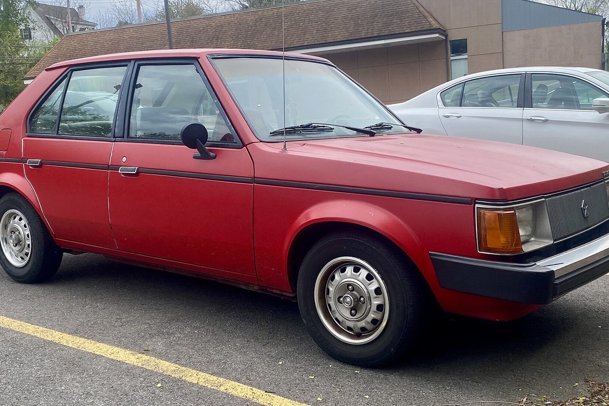 A red Dodge Omni five-door hatchback parked in a lot with yellow lines, seen from the front right