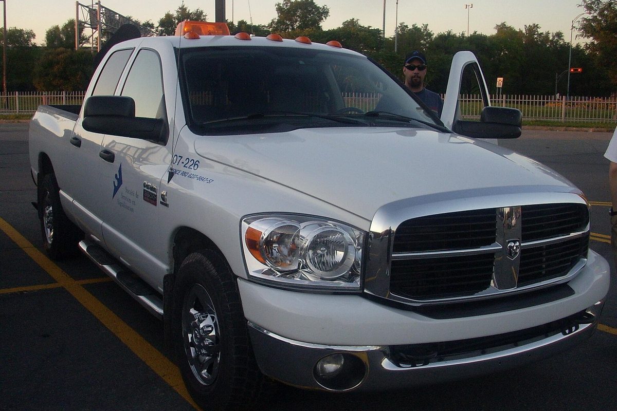 A white 2006-2008 Dodge Ram 3500 crew cab pickup with fleet decals and roof lights, driver's door open, in a parking lot at dusk