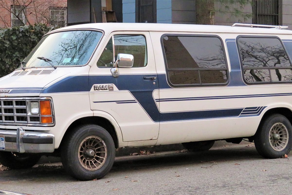 A white second-generation Dodge Ram Van with blue side stripes, side windows and a roof rack, parked on a city street