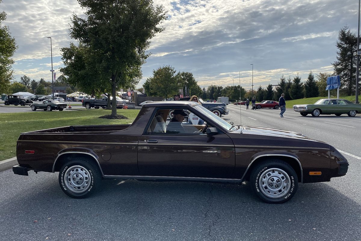 A dark brown Dodge Rampage car-based pickup with a short cargo bed behind the cab, seen from the side on a show-field road with other old cars behind it