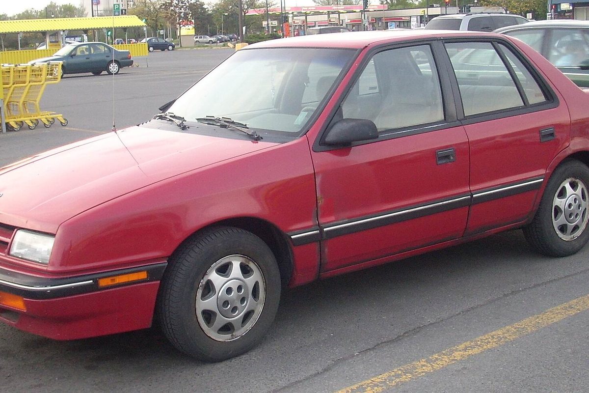A faded red Dodge Shadow five-door hatchback with gray side trim, parked in a store parking lot
