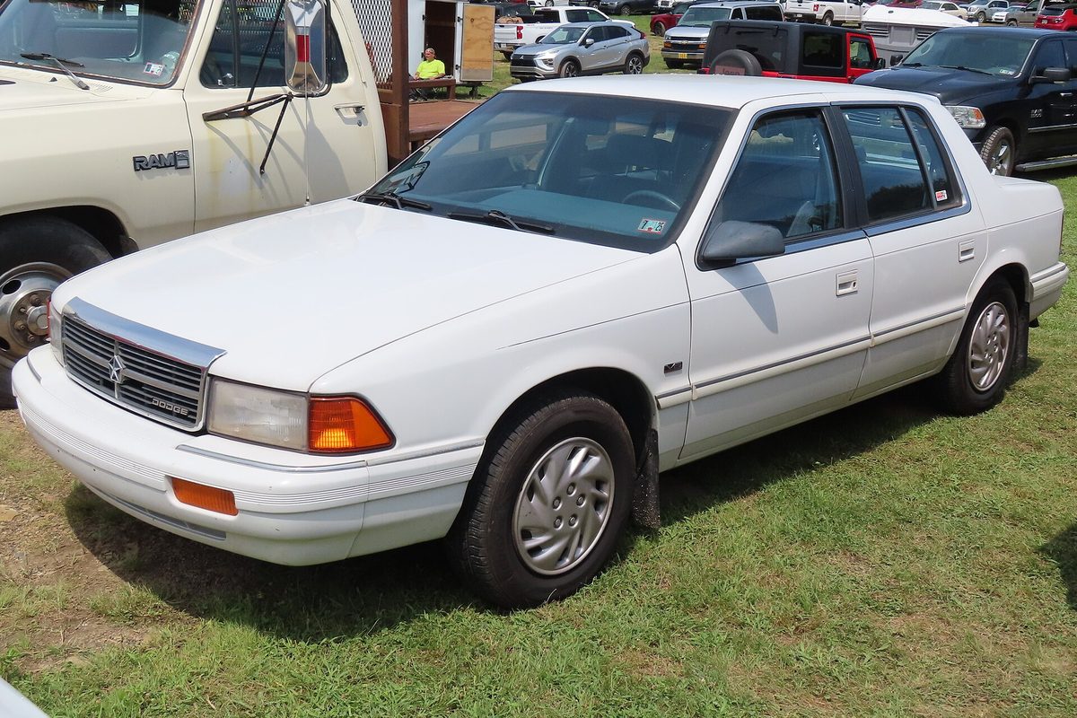 A white Dodge Spirit sedan parked on the grass at an outdoor car show, next to an older Dodge pickup