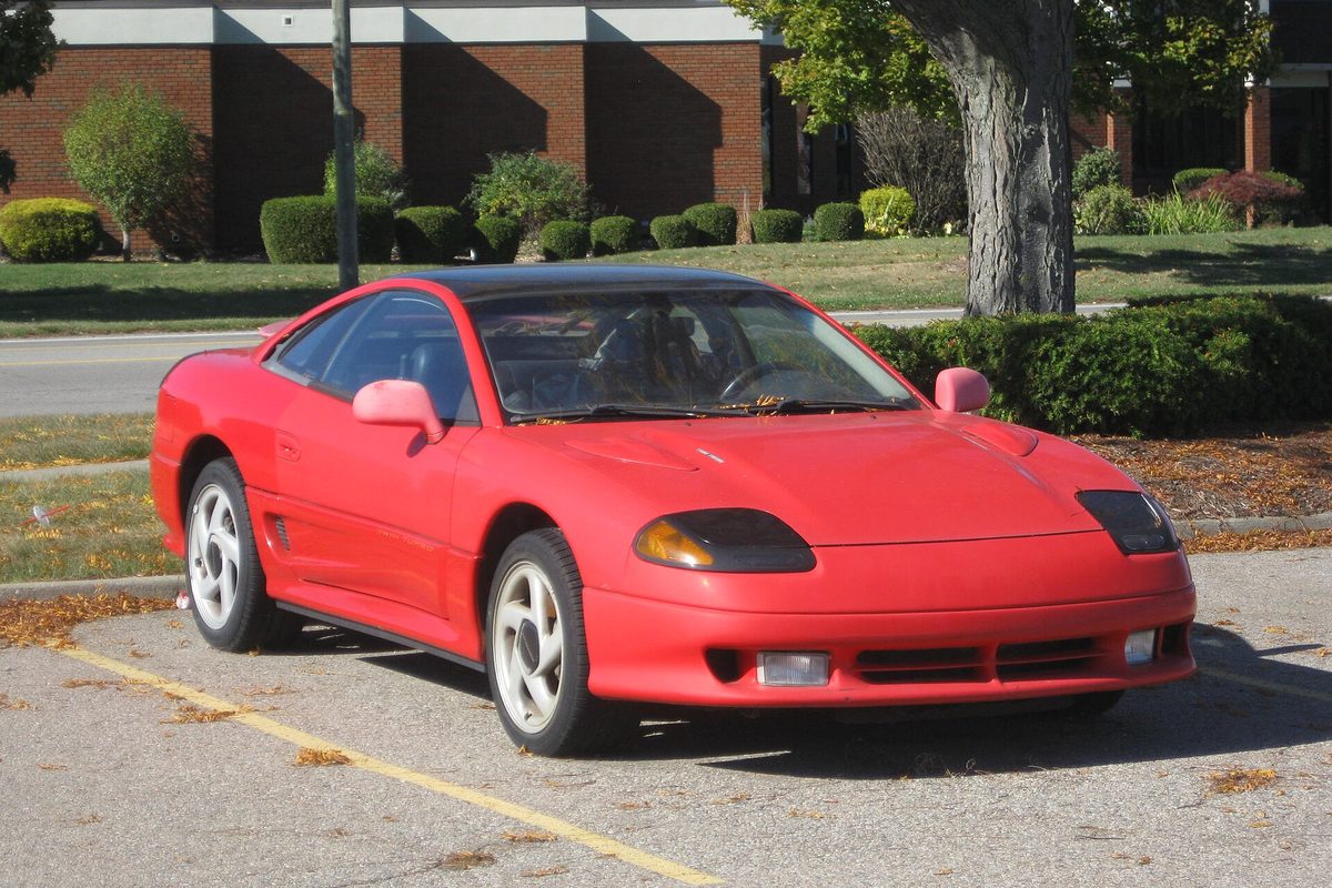 A red Dodge Stealth two-door liftback coupe with white five-spoke wheels and a dark roof, parked in a lot beside a lawn