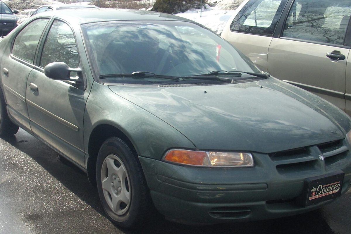 A dark green 1997-1998 Dodge Stratus sedan with steel wheels parked beside a minivan, snowbanks behind it