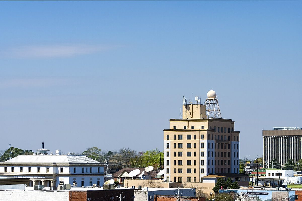 Downtown Dothan rooftops under a blue sky, with a tan mid-rise tower topped by a white sphere on a lattice mast and a white columned building to the left