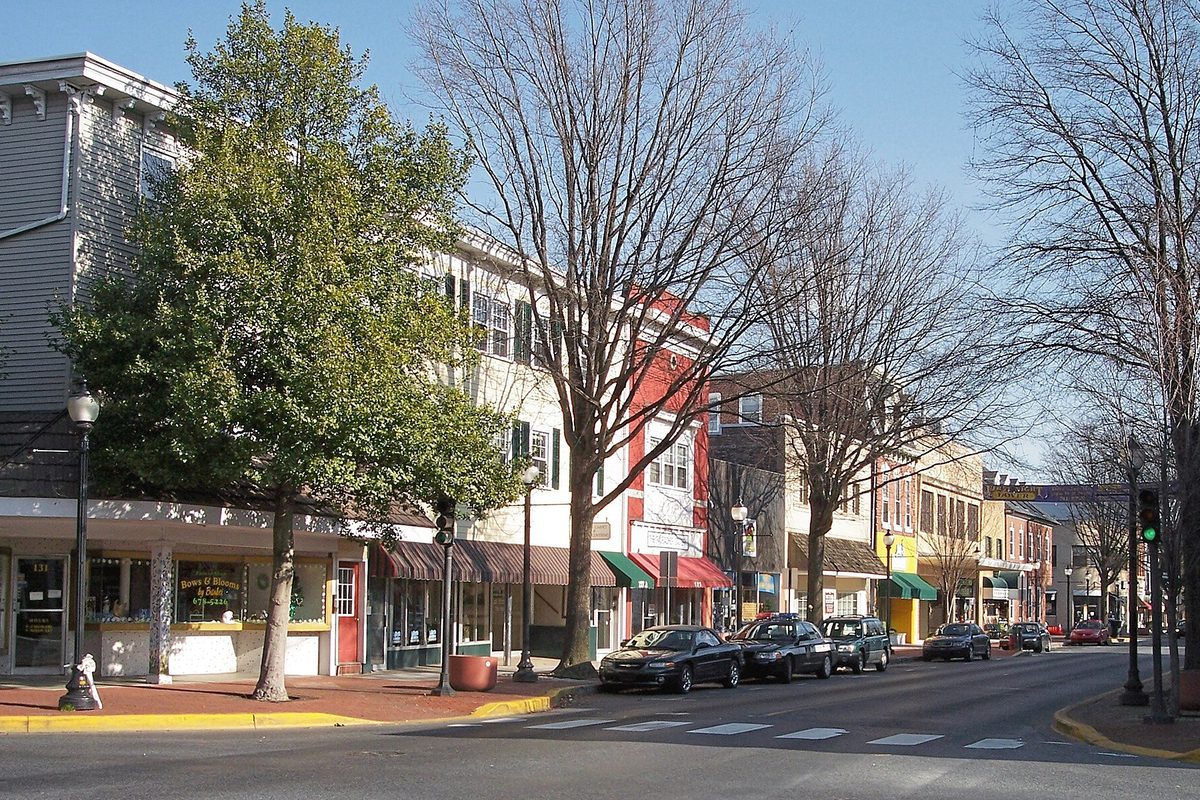 West Loockerman Street in downtown Dover on a sunny day, with two- and three-story storefronts, striped awnings, bare trees and cars parked at the curb