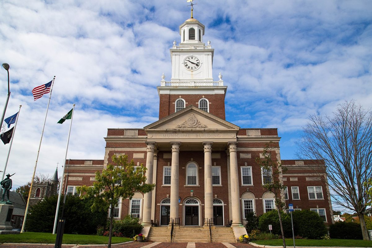 Dover City Hall, a red-brick building with a white-columned entrance and a white clock tower, flags flying out front under a partly cloudy sky