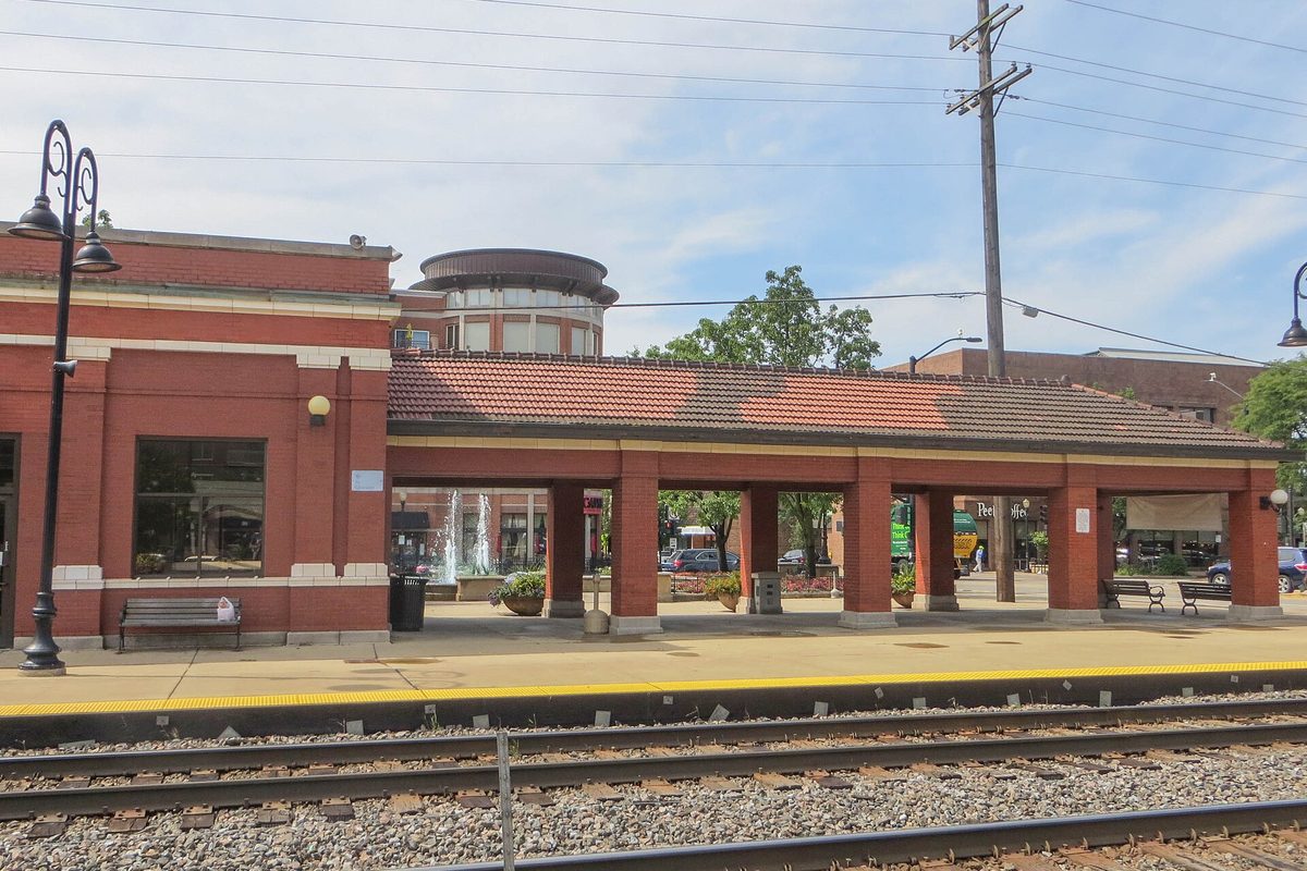 The Downers Grove Metra station, a red brick building with a covered colonnade and tile roof beside the platform and tracks on a sunny day