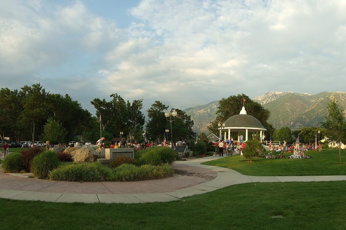 A white gazebo on the lawn of Draper Historic Park with a crowd gathered around it and the Wasatch Mountains behind
