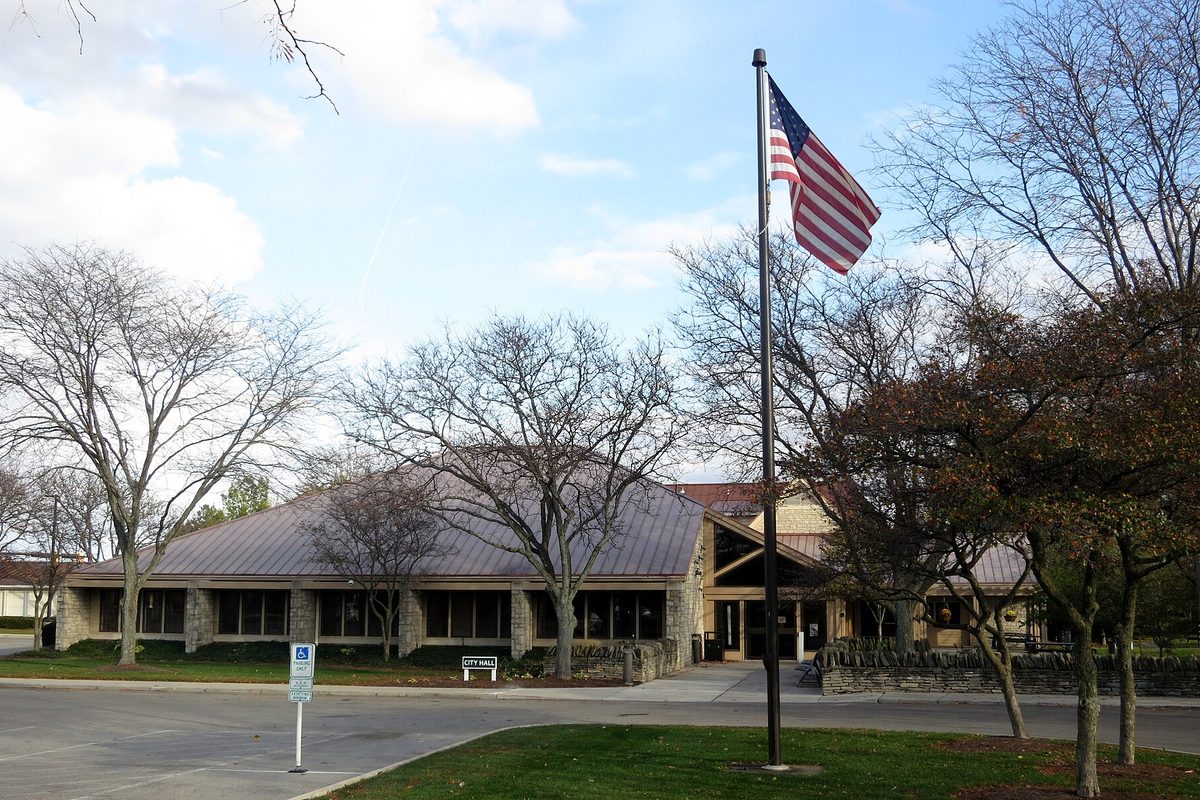 Dublin City Hall on Emerald Parkway, a low stone building with a sloped metal roof, seen from the parking lot past a flagpole with the U.S. flag and bare trees