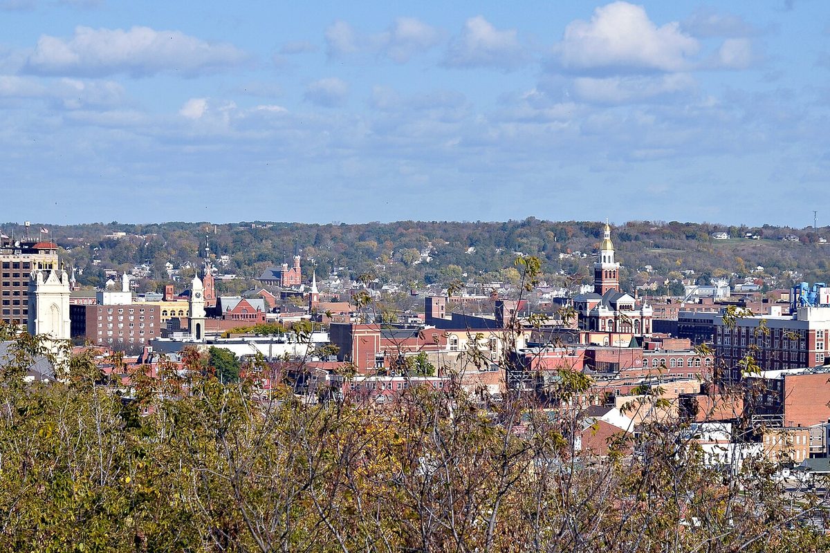Downtown Dubuque seen from a bluff, with brick buildings, church steeples and a gold-domed clock tower in front of tree-covered hills