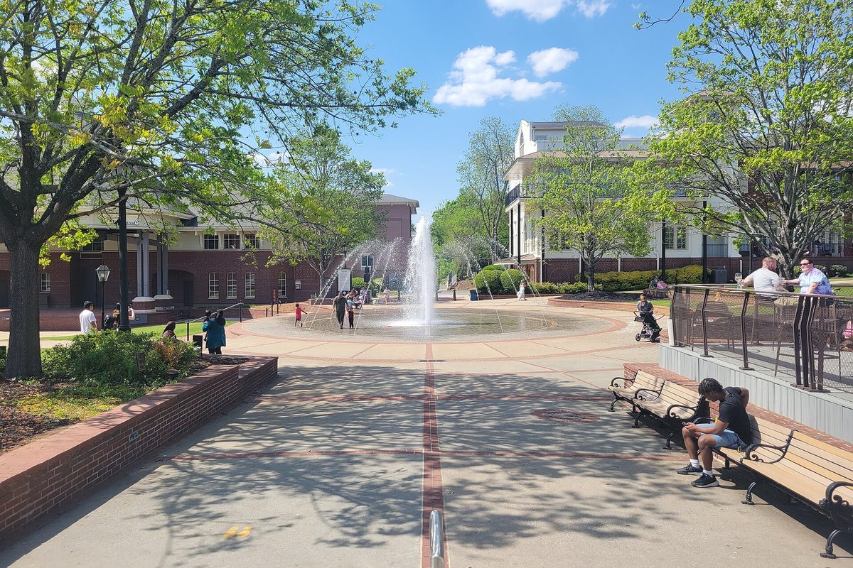A fountain in the middle of a brick-paved plaza in Duluth, with benches, spring trees, people strolling and brick buildings behind