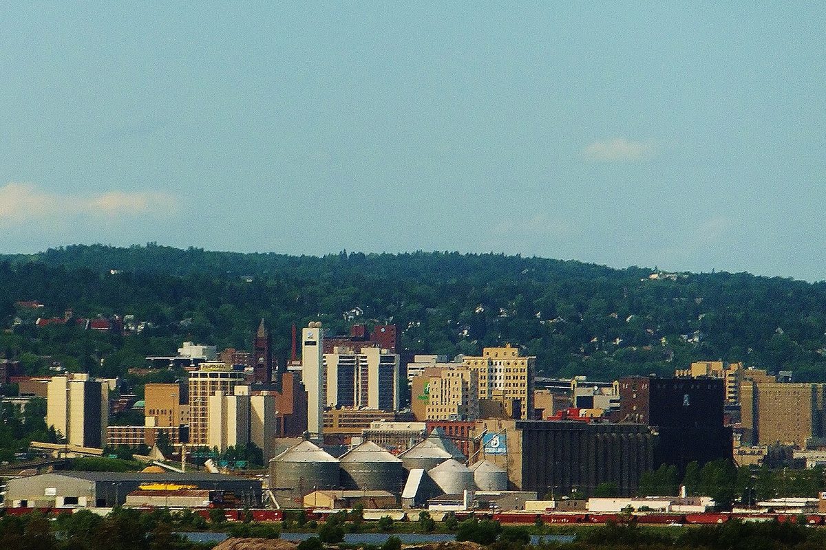 Downtown Duluth's office towers and grain elevators along the waterfront, with a wooded hillside rising behind the city