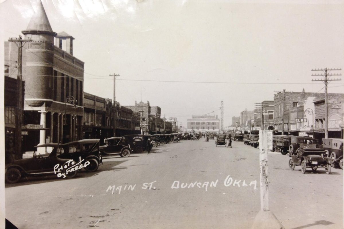 A 1920s black-and-white photo of Main Street in Duncan, with early cars parked along both curbs and a brick building with a round turret on the left