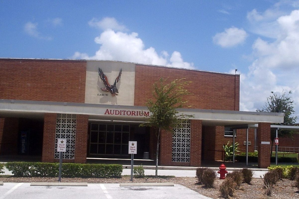 The brick auditorium wing of Dunedin High School, with an eagle emblem above the entrance, palms and shrubs out front and a blue sky