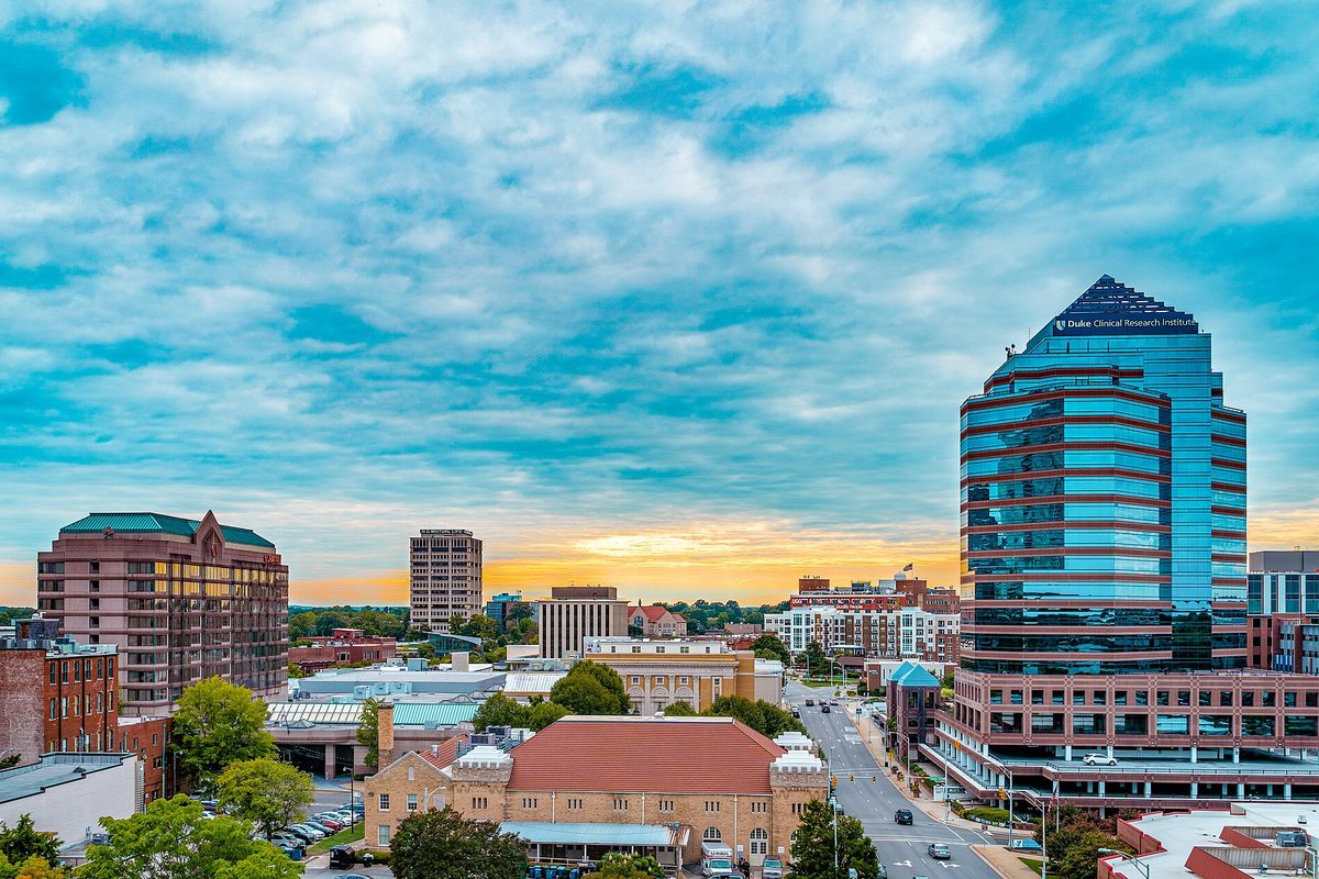 Downtown Durham's skyline at sunset under streaks of blue cloud, with a tall glass office tower on the right and brick buildings below