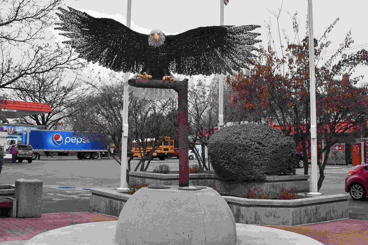A bronze eagle sculpture with spread wings on a pillar in a small plaza in Eagle, with a delivery truck and a school bus behind