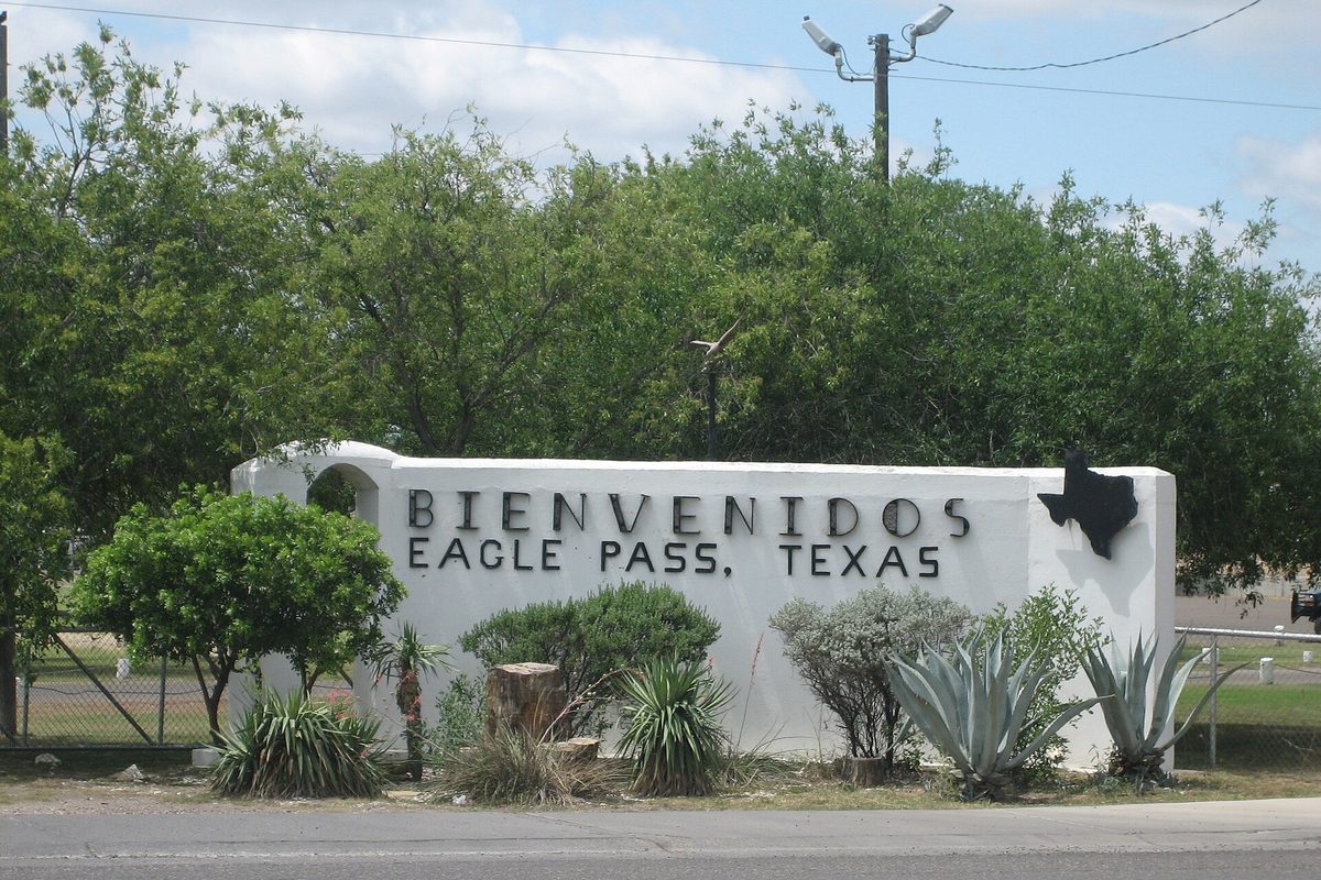 A white stucco welcome wall reading Bienvenidos Eagle Pass, Texas, with a black outline of Texas, agave and shrubs in front and a row of trees behind