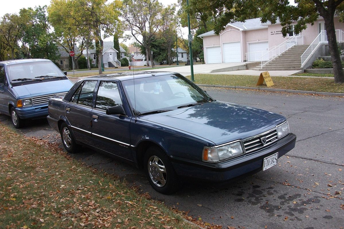 A dark blue Eagle Premier sedan parked at a curb on a leafy residential street, a blue van behind it