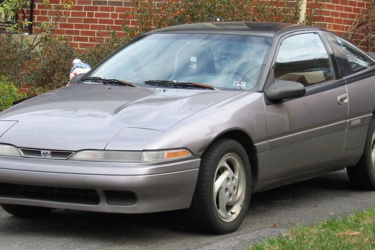 A gray first-generation Eagle Talon hatchback coupe parked on a driveway beside a brick wall