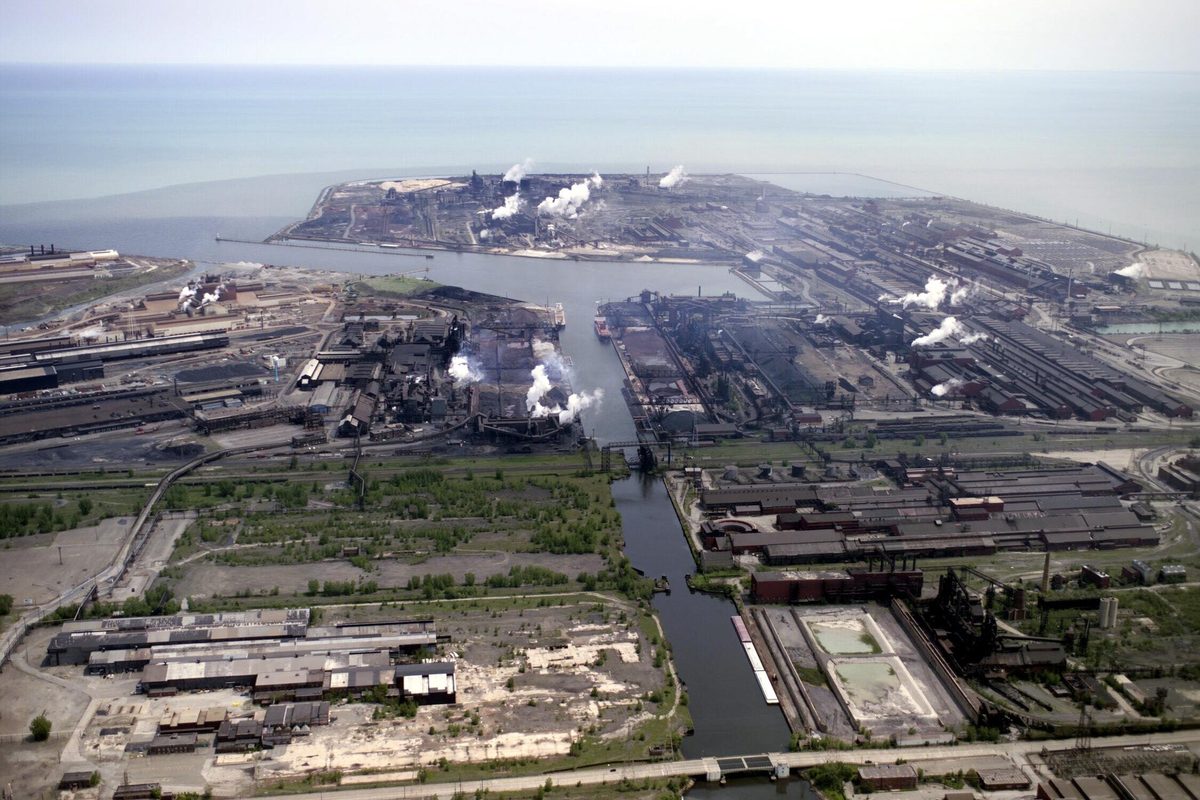 Aerial view of the Indiana Harbor and Ship Canal in East Chicago, lined with steel mills and white smoke plumes, with Lake Michigan behind