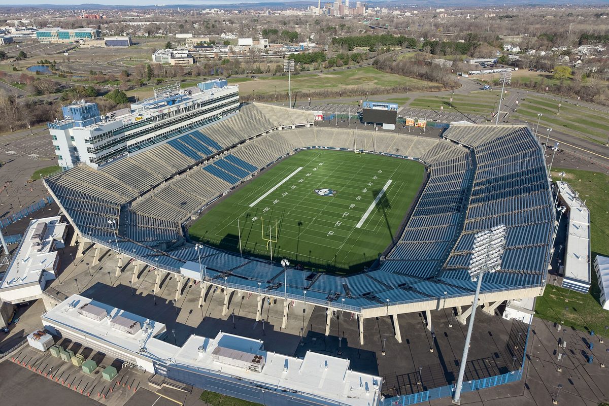 Aerial view of Pratt and Whitney Stadium at Rentschler Field in East Hartford, its green football field ringed by blue seats, with the Hartford skyline in the distance
