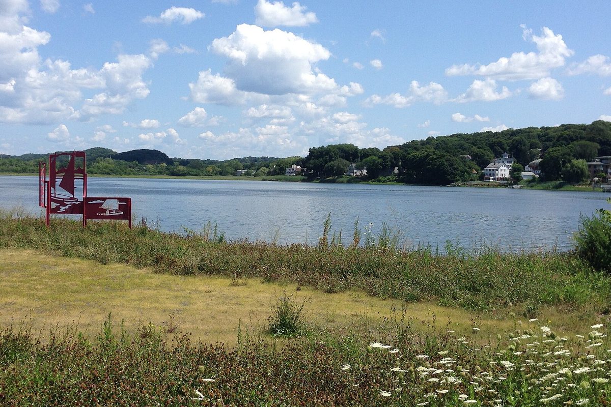 The Quinnipiac River seen from the Fair Haven side in New Haven, with a red metal sailboat sculpture on the grassy bank and wooded hills across the water