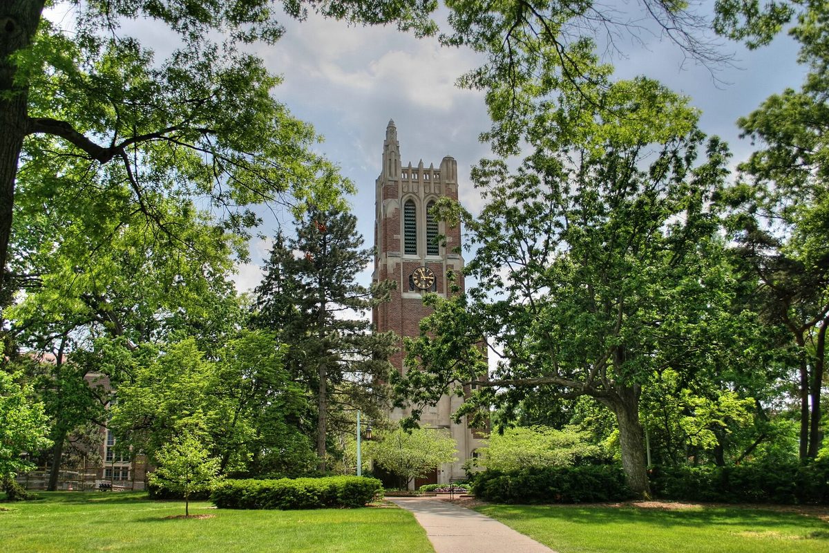 Beaumont Tower, a red brick bell tower with a clock face, framed by tall green trees and a lawn on the Michigan State University campus