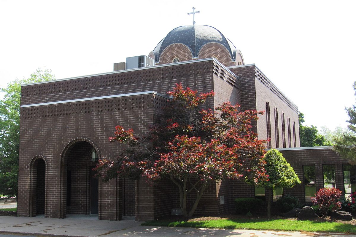 The brown brick Assumption Greek Orthodox Church in East Moline, with a dark dome and cross on top and a red-leafed maple in front
