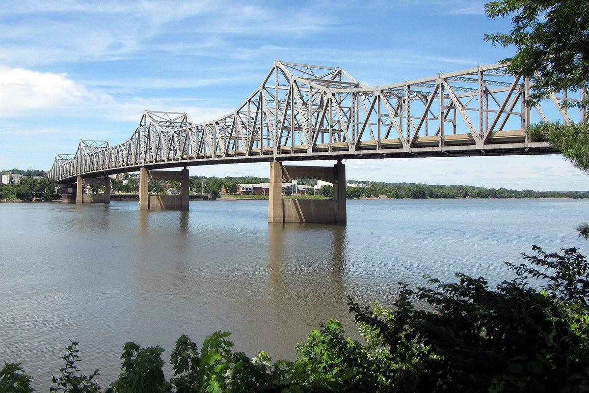 The steel truss Murray Baker Bridge crossing the wide Illinois River, seen from the East Peoria bank under a blue sky with green brush in front