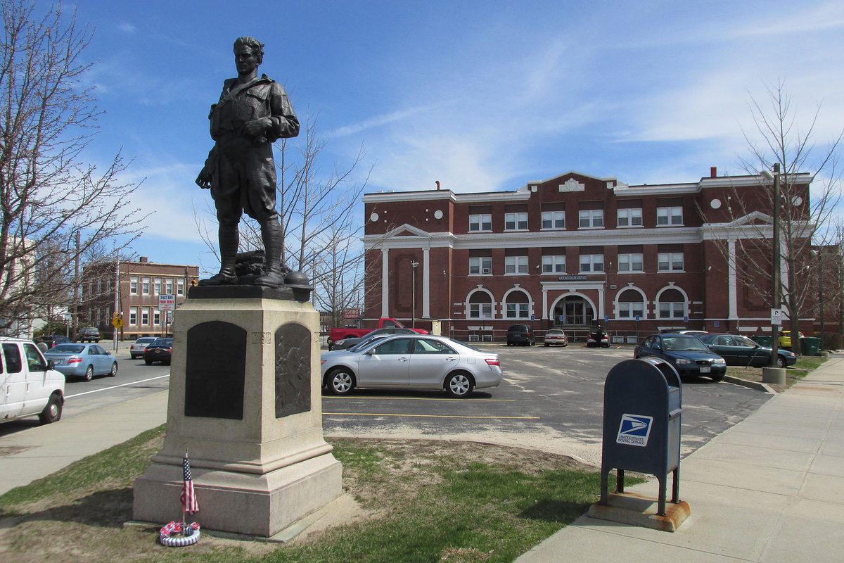 A bronze World War I soldier statue on a stone pedestal at Taunton Plaza in East Providence, with a red brick building, parked cars and a blue mailbox behind it