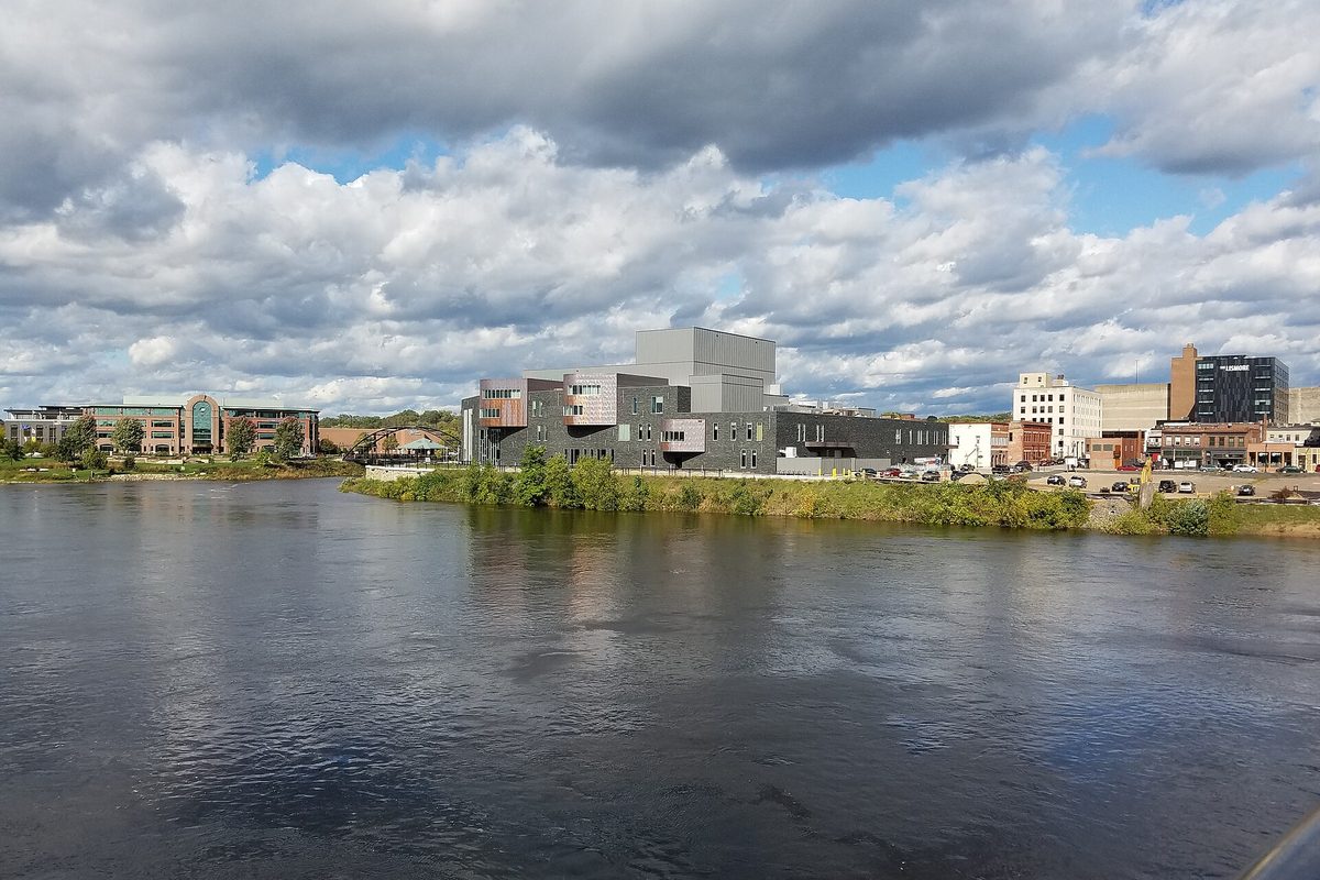 Downtown Eau Claire seen across a wide river, with a large modern gray building on the grassy bank and taller downtown blocks behind it under heavy white clouds