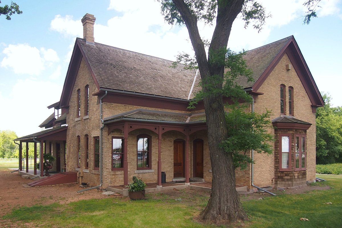 The yellow brick John R. Cummins Farmhouse in Eden Prairie's Silverness Park, with a tall tree and red trim on the porch