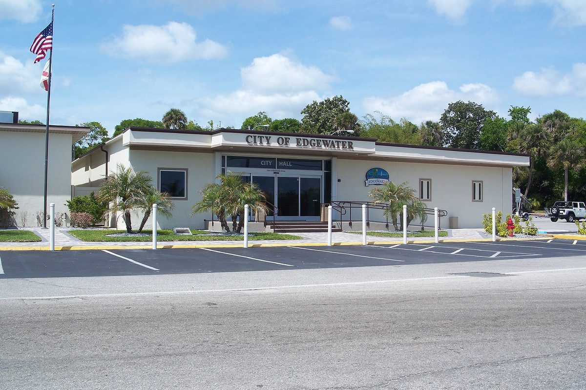 Edgewater City Hall, a low cream-colored building with City of Edgewater lettered above the doors, an American flag, palm trees and an empty parking lot