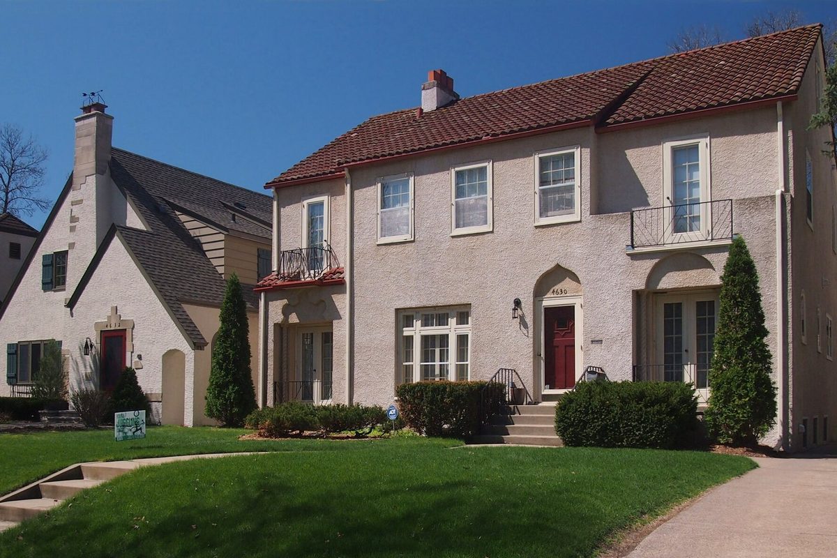 Two late-1920s houses on Casco Avenue in Edina's Country Club district, a stucco one with a red tile roof and a Tudor-style one beside it, behind clipped hedges and a green lawn