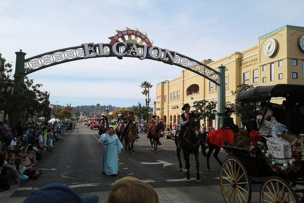 The Downtown El Cajon arch over Main Street during a parade, with a woman in a long blue costume, riders on horseback and a horse-drawn carriage passing crowds