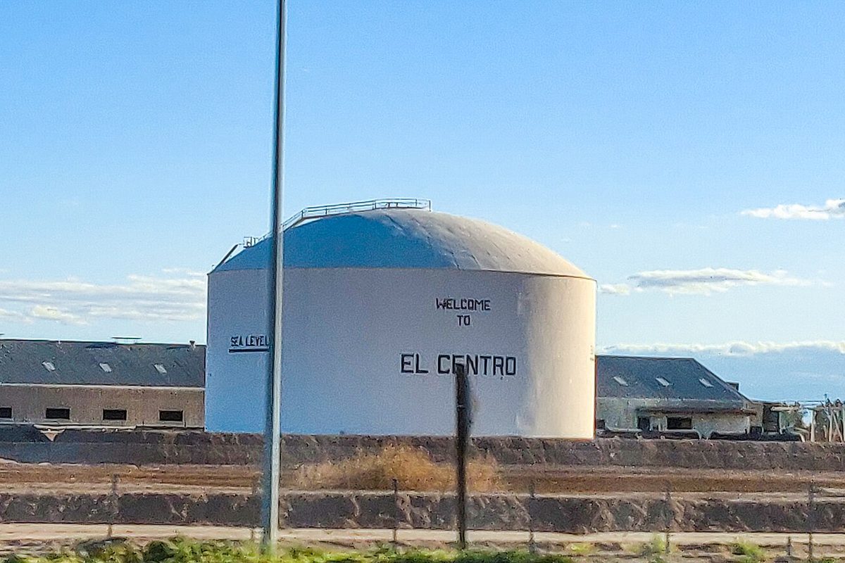 A white domed water tank painted Welcome to El Centro, with a small Sea Level sign, beside low buildings under a blue sky