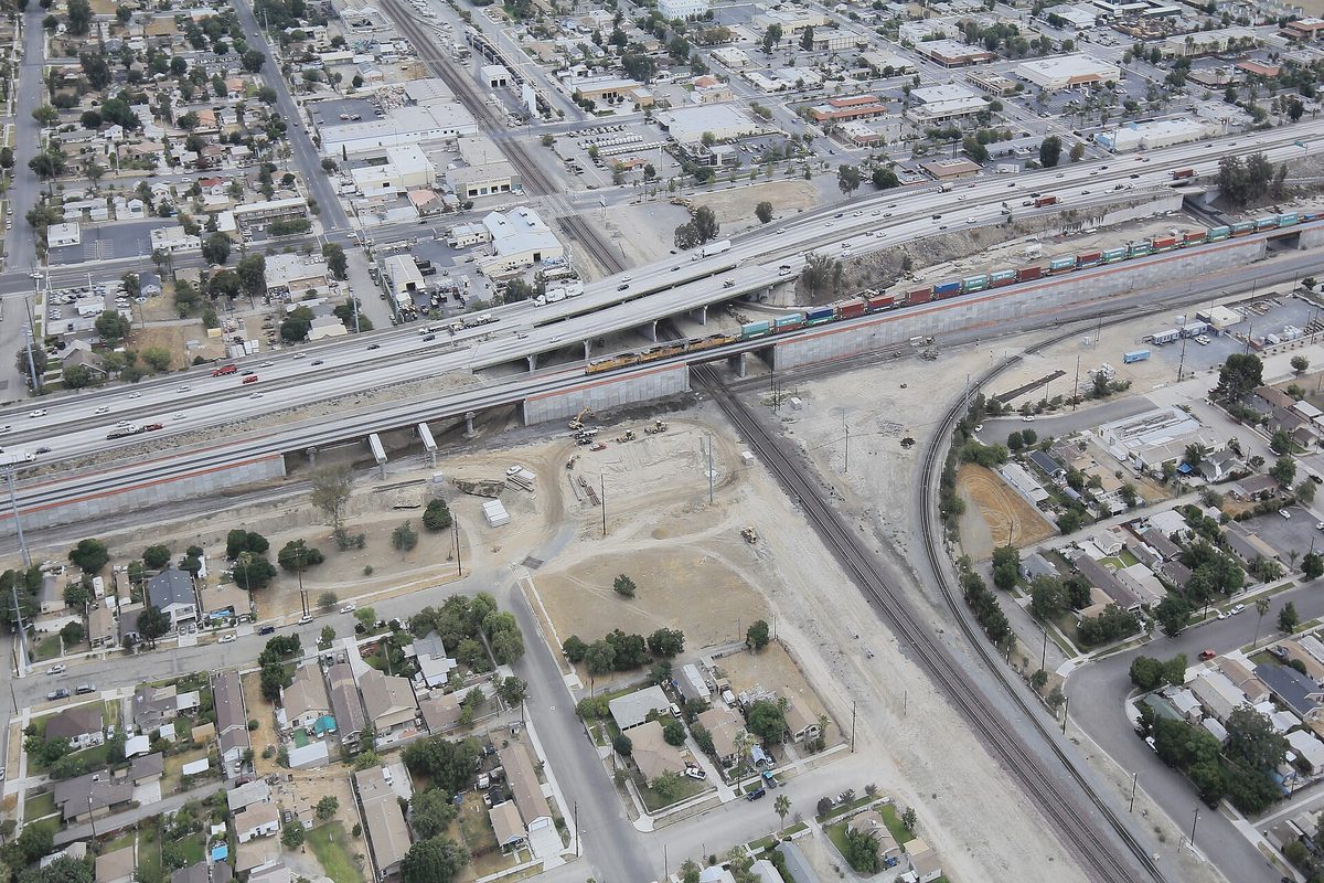 Aerial view of a freeway and a railroad crossing over each other on a grade separation, with a freight train, rows of houses and bare dirt lots below