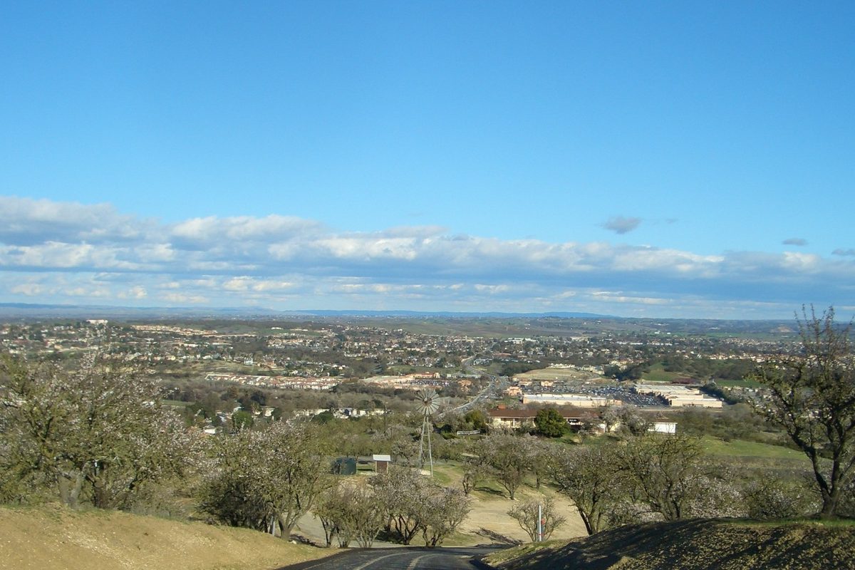 Hilltop view across Paso Robles toward a wide valley under a blue sky with white clouds, with bare trees and a dry grassy slope in front