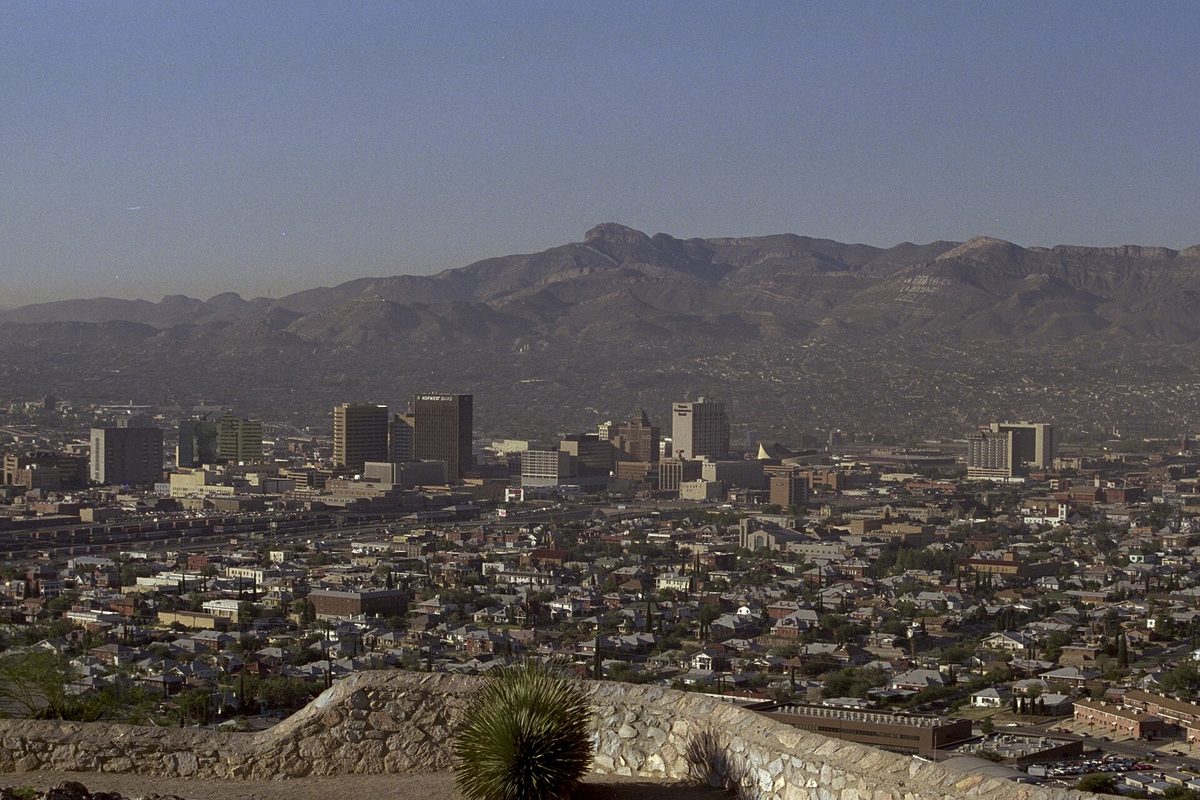 Downtown El Paso's towers and miles of low-rise neighborhoods seen from a hillside overlook, with brown mountains on the horizon and a yucca in the foreground