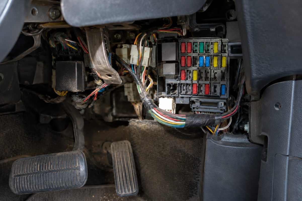 An open panel under a car's dashboard showing a fuse box of colored fuses and taped wiring bundles above the brake and gas pedals