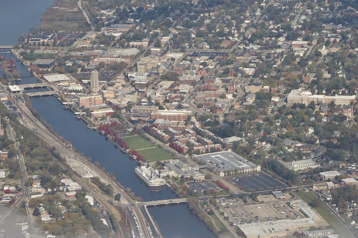 Aerial view of downtown Elgin on the Fox River, with a large white boat docked by a green sports field, parking lots, and blocks of homes and church spires behind