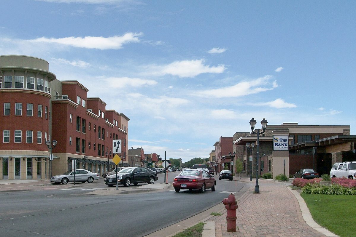 Main Street in downtown Elk River, with red-brick buildings and a rounded corner tower on the left, a red sedan driving past, a fire hydrant on a brick sidewalk and a bank sign on the right